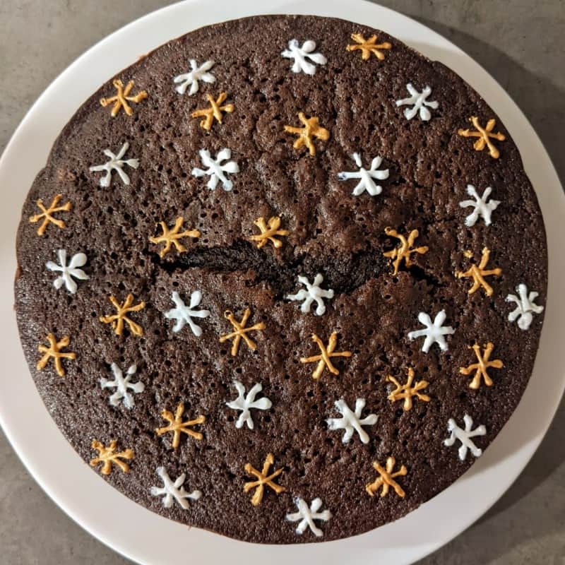 A round, dark brown chocolate cake seen from above. The top of the cake shows some gently bumpy texture. It is decorated with gold and silver piped stars or sparkles.