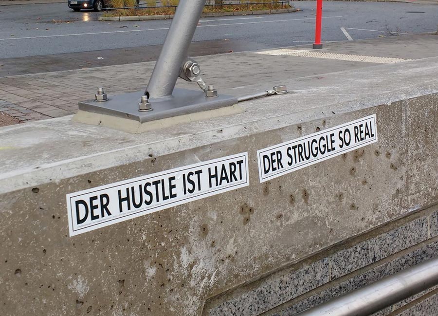 A wall with stickers reading DER HUSTLE IST HART and DER STRUGGLE SO REAL